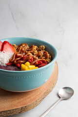 acai bowl with strawberries, granola, mango, coconut, peanut butter , green marble background, wood plate, blue bowl.
