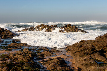 Beautiful coastline of the Atlantic Ocean with waves crashing against brown rocks, splashing water and spreading white foam, Portugal