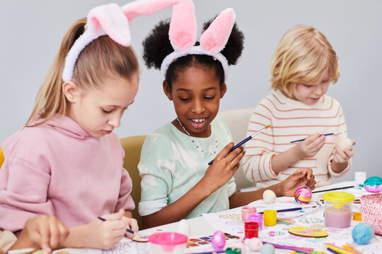 Diverse group of happy children painting Easter eggs and wearing bunny ears while enjoying art and craft class in school