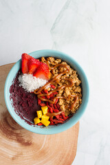 acai bowl with strawberries, granola, mango, coconut, peanut butter , green marble background, wood plate, blue bowl.