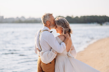 Senior newlyweds hug. Stylish couple of elderly newlyweds stand embracing on river bank.