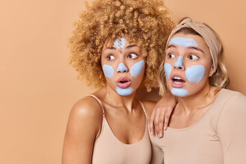Horizontal shot of shocked amazed women stare away gasp from surprise cannot believe in something terrifying apply beauty mask wear casual clothes isolated over beige background blank space.