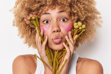 Horizontal shot of curly haired woman keeps lips rounded applies pik hydrogel patches under eyes holds plants over face uses natural products for making cosmetics stands bare shoulders indoor