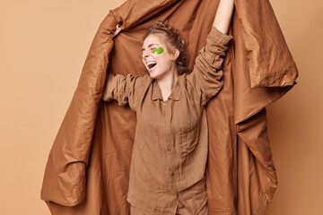 Good morning concept. Relaxed young woman holds blanket yawns and stretches arms awakes after sleeping applies hydrogel beauty patches dressed in comfortable pajama isolated over brown background