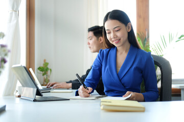 Asian business woman sitting at her office desk and making note on notebook.