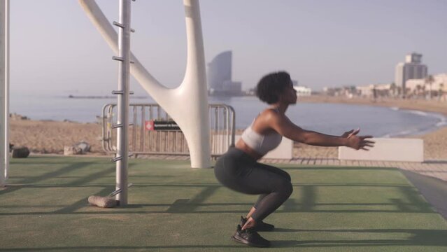 Black afro American fitness woman workout squat sequence exercise in open air gym on sandy beach with cityscape of Barcelona city on background.