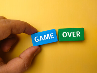 Top view hand holding colored block with text GAME OVER on a yellow background.