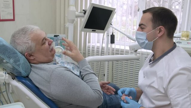 Elderly man sitting in dentist's chair talks about his dental problems. Male dentist in dental office talking with senior patient and preparing for treatment. Healthcare, medicine and dentistry