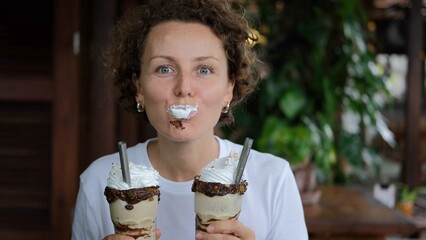 Funny curly young woman playing, enjoying, eating whipped cream from two glasses of chocolate milk...