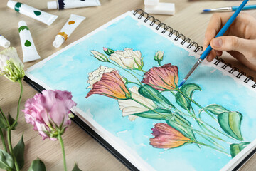 Woman painting eustomas in sketchbook at wooden table, closeup