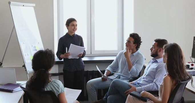 Diverse businesspeople gather in office boardroom take part in corporate training led by confident Indian woman coach speaking standing near flip chart in front of staff. Seminar, teambuilding concept