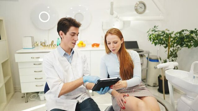Young male dentist and female patient looking at digital tablet, discussing dental treatment plan