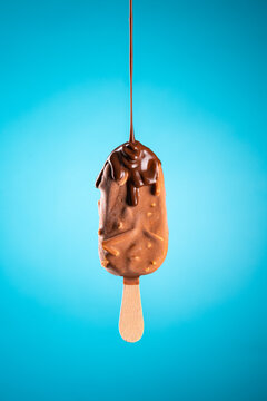 Chocolate popsicle ice cream with almond nuts flavour on wood stick and liquid chocolate falling, isolated on blue background.