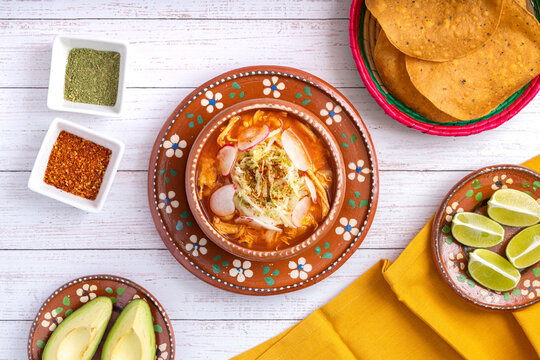 Traditional mexican food. Red pozole soup with chicken accompanied with avocado, lettuce, onion, radish, lemon, chili and crispy corn tortillas also known as tostadas on a white wooden background.