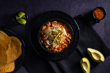 Traditional mexican food. Red pozole soup with chicken accompanied with avocado, lettuce, onion, radish, lemon, chili and crispy corn tortillas also known as tostadas on a black wooden background.
