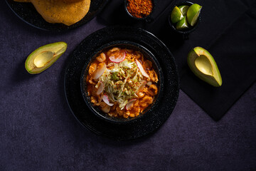 Traditional mexican food. Red pozole soup with chicken accompanied with avocado, lettuce, onion, radish, lemon, chili and crispy corn tortillas also known as tostadas on a black wooden background.