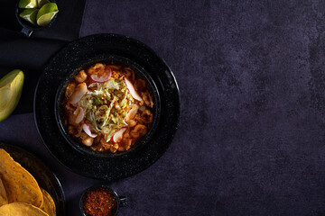 Traditional mexican food. Red pozole soup with chicken accompanied with avocado, lettuce, onion, radish, lemon, chili and crispy corn tortillas also known as tostadas on a black wooden background.