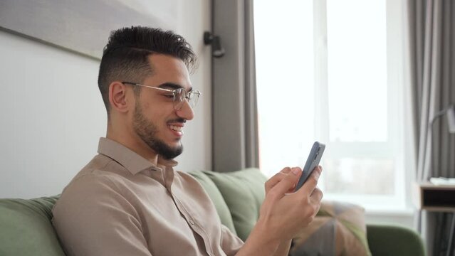 Happy young man smiling looking at cell phone sitting in living room at home