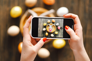 Woman holding mobile phone and making photo of eggs painted for Easter isolated on wooden...