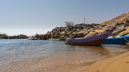 Fototapeta premium Old painted wooden boats are moored at the sandy shore of the Nile. Ripples on clear water. Picturesque boulders on the shore. Blue sky. Egypt