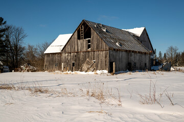 Fototapeta na wymiar An aging wooden pole barn slowly collapsing. Shot on a sunny day in winter with fresh snow and blue sky.