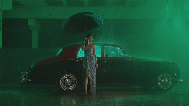 Beautiful Elegant Young Woman Standing and holding umbrella near old retro classic car. Girl with umbrella stands in street under the rain . Old fashioned luxury style concept . Autumn rainy day