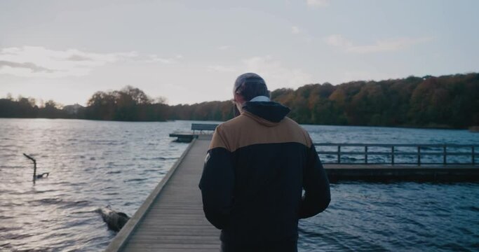 Young Tall Scandinavian Guy Walks Towards a Bench On a Bridge by Gyllebo Lake in Autumn Sunset - Tracking Medium Shot From behind