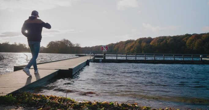 Young Scandinavian Man Walks On a Wooden Bridge By Gyllebo Lake and Sits Down on a Bench - Static Wide Shot