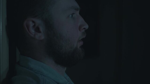 A young man with a light beard speedily runs around a corner and presses his back to the wall, out of breath as if someone was chasing him into the dimly lit house