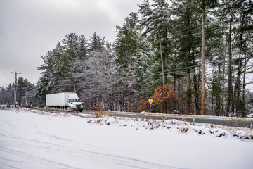 Day cab big rig semi truck transporting cargo in dry van semi trailer running on the local road along the forest with snow and ice at winter season