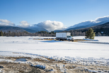 Day cab middle power rig white semi truck with long box trailer delivered cargo on the place on big meadow covered with snow and surrounded winter trees and mountain.