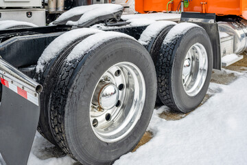 Drive axels of orange big rig semi truck with double wheels and tires on the winding parking lot with snow and ice