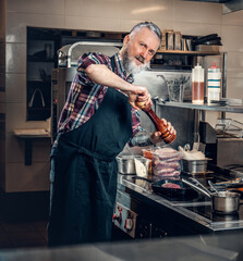 Old man chef peppering meat looking at camera in kitchen