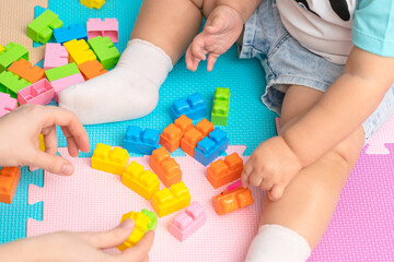 Toddler sits on the mat in the playroom and learns with the help of his mother to assemble the bricks of a plastic constructor.