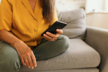 Close-up picture of hands of elderly woman holding smartphone sitting on grey couch in yellow blouse and olive green jeans, wearing silver ring on finger, using gadget to surf Internet