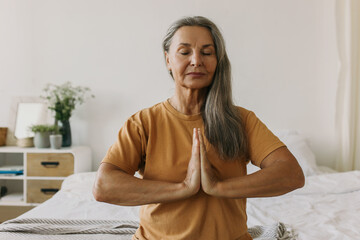 Human, wellness, well-being, sport. Indoor picture of charming retired lady of 60s in yellow t-shirt sitting on bed with eyes closed, keeping hands in namaste gesture or praying position - obrazy, fototapety, plakaty
