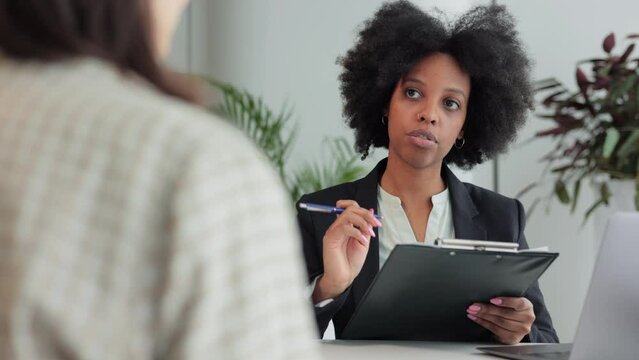 Female employer interviewing job seeker and sitting at table with device in modern office spbi. 4k video African american businesswoman talks to female candidate and holds resume, looks with smile and