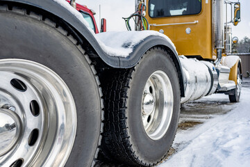 Yellow professional day cab big rig semi truck with twin axles with wheels and tires with a large tread standing on the winter parking lot with snow and ice - obrazy, fototapety, plakaty