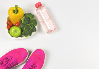 vegetables  capsicum, broccoli, tomatoes and apple  in heart shape plate,  pink sneakers or sport shoes and bottle of water  on white  background with copy space. healthy lifestyle.