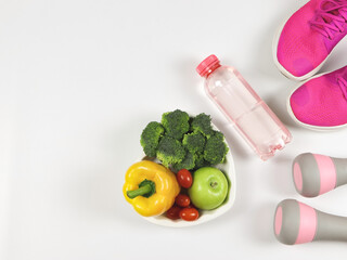 capsicum, broccoli, tomatoes and apple  in heart shape plate,  pink gray dumbbells, pink sneakers or sport shoes and bottle of water  on white  background with copy space. healthy lifestyle.