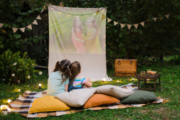 Open air cinema. Backyard Family outdoor movie night with kids. Sisters spending time together and watching cimema at backyard. DIY Screen with film. Summer outdoor weekend activities with children