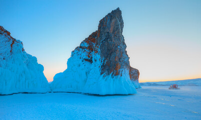 A granite rock with steep slopes rises above a frozen lake with reflection on the ice at sunset -  Baikal lake © muratart
