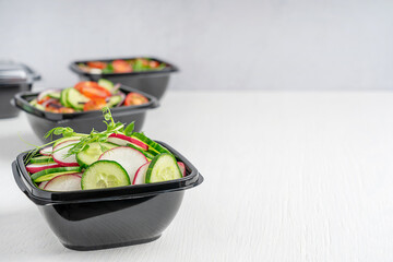 Vegan salads of cucumber slices, radish and tomatoes served in disposable black plastic bowls used...