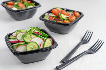 Vegetarian salads of cucumber slices, radish and tomatoes served in disposable black plastic bowls used for food delivery served with forks on white wooden background. Healthy eating concept