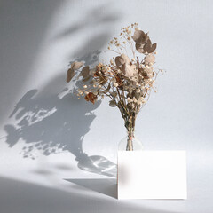 Blank white greeting card or note in front of bouquet of dried flowers in a vase, with shadow. Minimalist mock up template.
