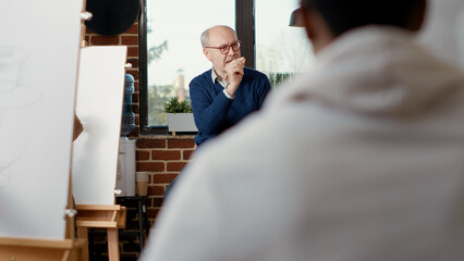 Senior man teaching art students to draw artwork on canvas, attending creative workshop to explain drawing practice technique. Educational program to learn new artistic skills, creating sketch.
