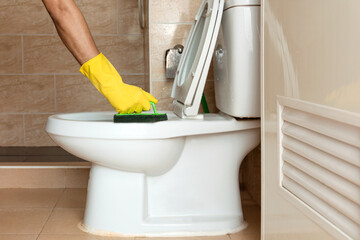 Human hand in a yellow rubber glove is using a plastic brush to scrub the toilet bowl.