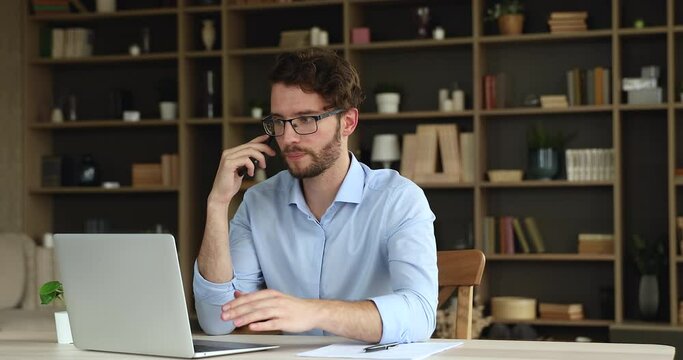 Confident millennial businessman sit by laptop negotiate with customer using phone discuss project details explain new service advantages. Young man specialist help client remotely give expert advice
