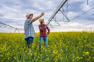 Two generations farmers are standing in their rapeseed field. Senior man is teaching his successor about the farming.