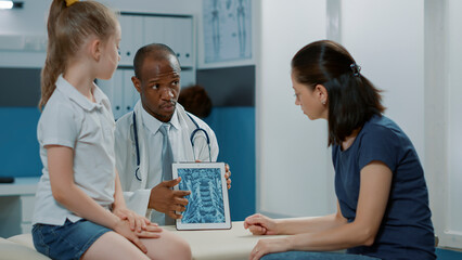 Specialist showing radiography diagnosis to mother and child at medical appointment. Physician explaining x ray scan results on digital tablet to help with treatment and recovery.